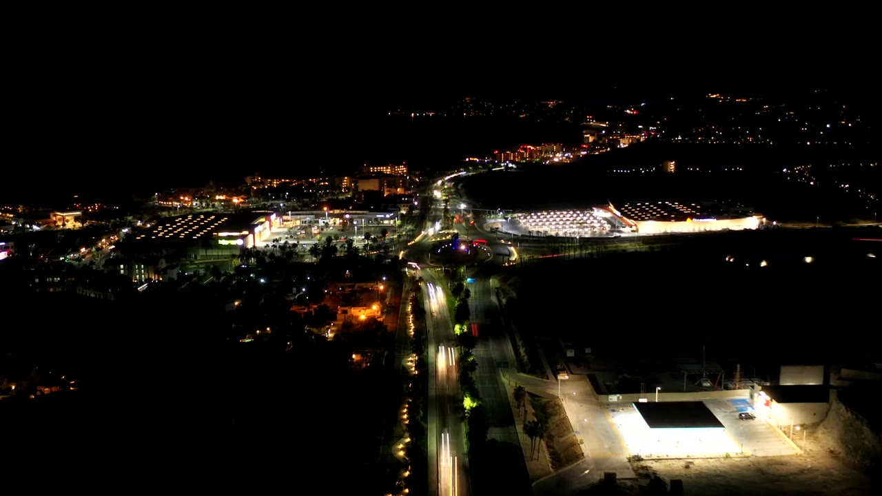 Los Cabos night drone view with the main road running toward a bright building complex, small traffic streaks, dark foreground land, and no visible people.