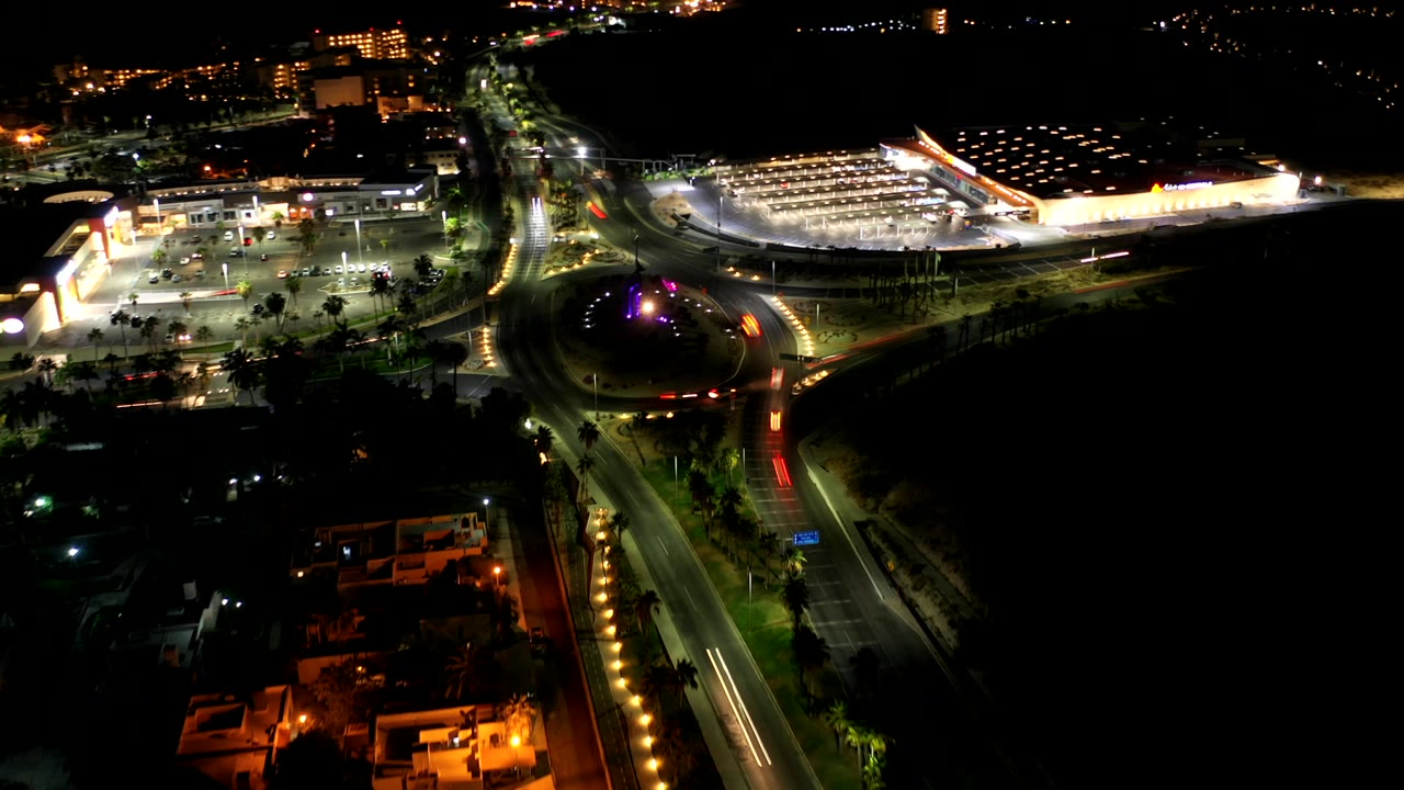 Night aerial view of a lit roundabout and roads beside a bright building complex.