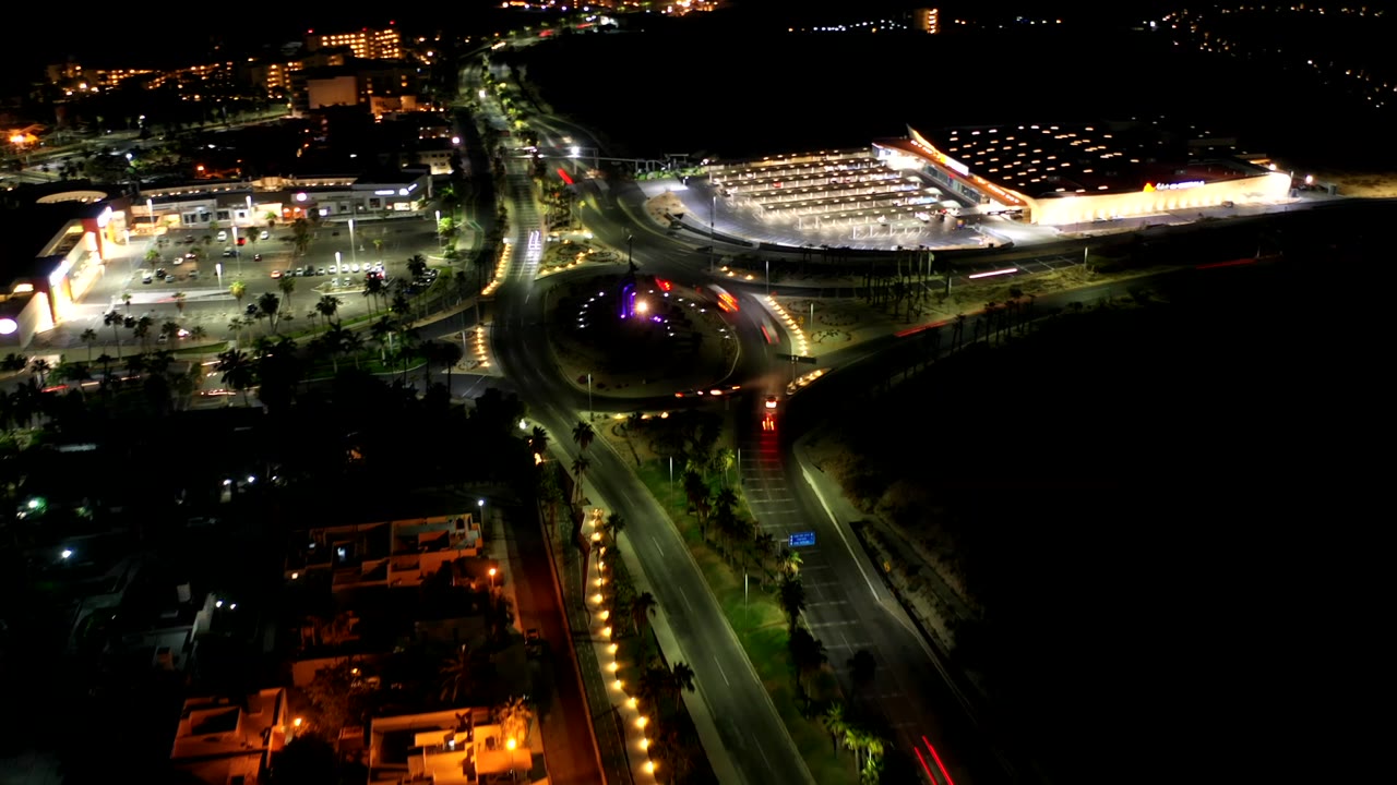 Los Cabos, Mexico aerial frame with the roundabout lower center, bright parking rows to the right, road light trails, palm medians, and no visible people.