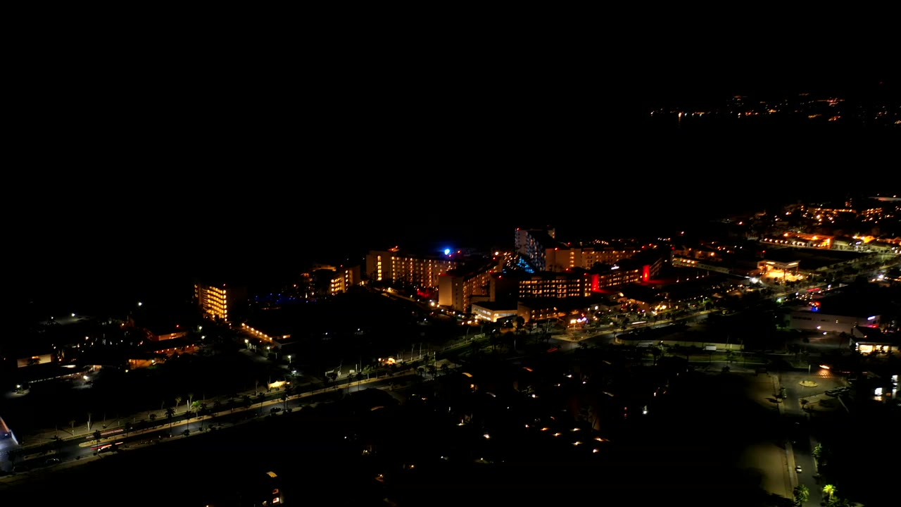 Opening night aerial view over Los Cabos with warm building lights, dark surrounding space, road lines, and no visible people.