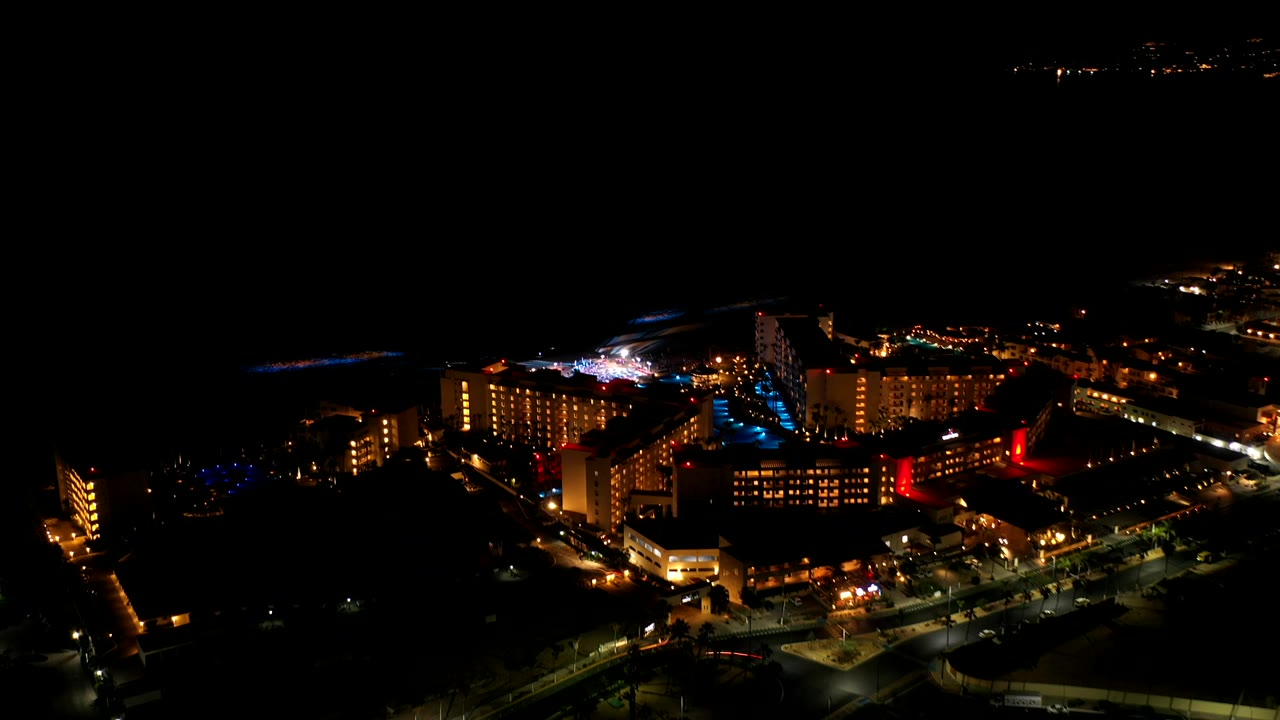 Night bird's-eye view over Los Cabos, Mexico, with lit hotel-like buildings, road lights, dark open space, and no visible people.