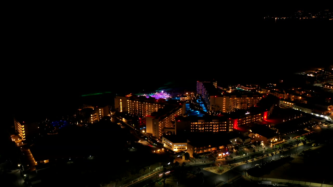 Los Cabos night aerial with a brighter cluster of buildings, blue pool lighting near the center, warm road lights, and no visible people.