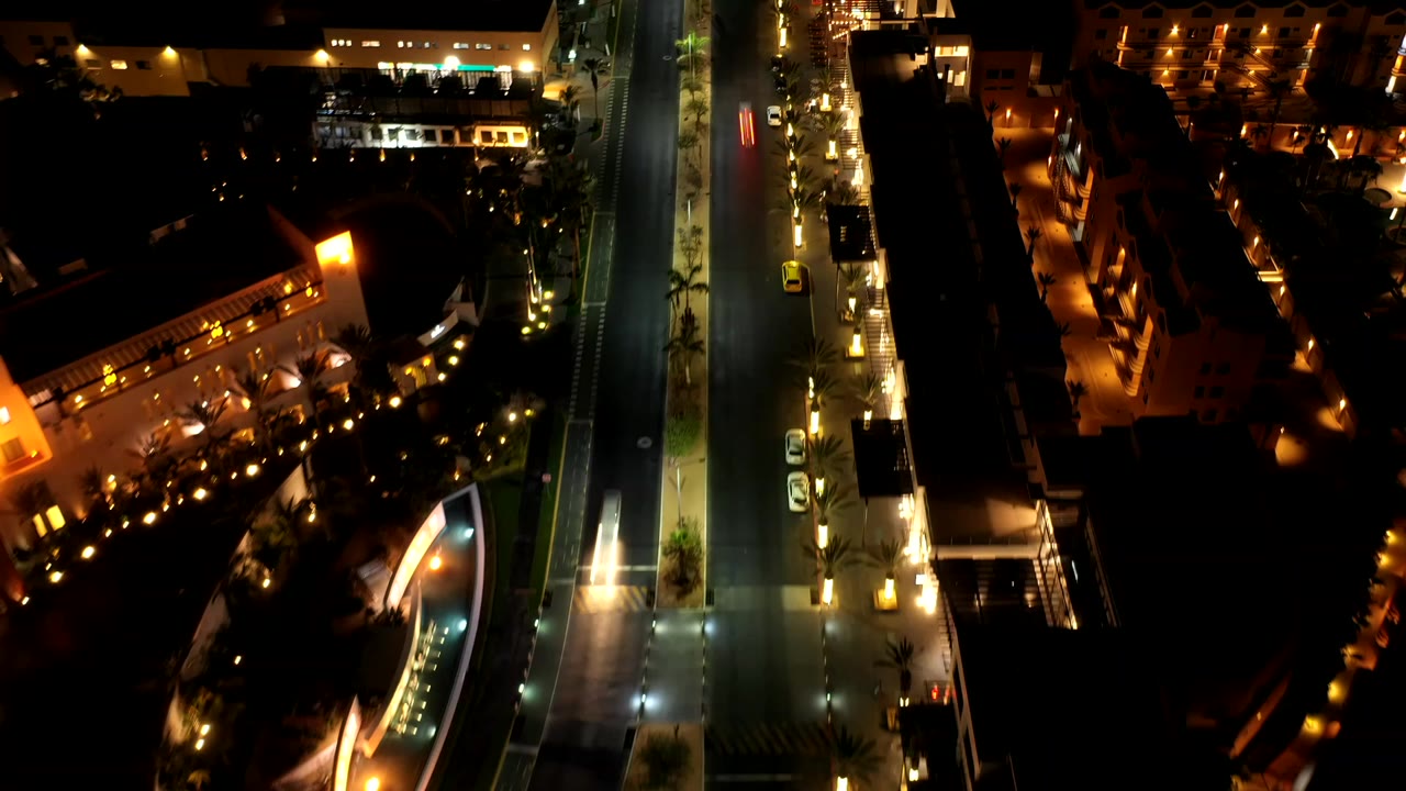 Night aerial frame over Los Cabos, Mexico, showing a vertical city road, warm building lights, palm-lined sidewalks, parked cars, dark rooftops, and no visible people.