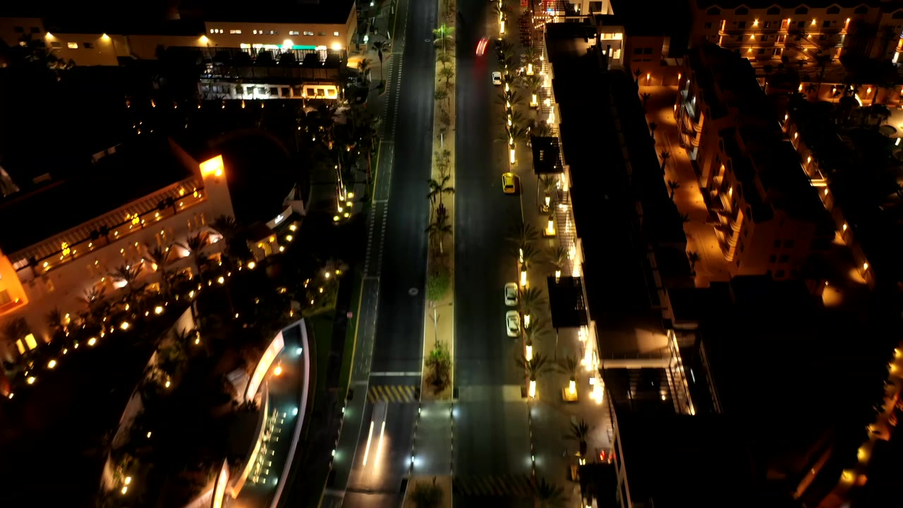 A high night view in Los Cabos, Mexico, centers on a bright road bordered by low buildings, rows of lights, parked vehicles, and no visible people.