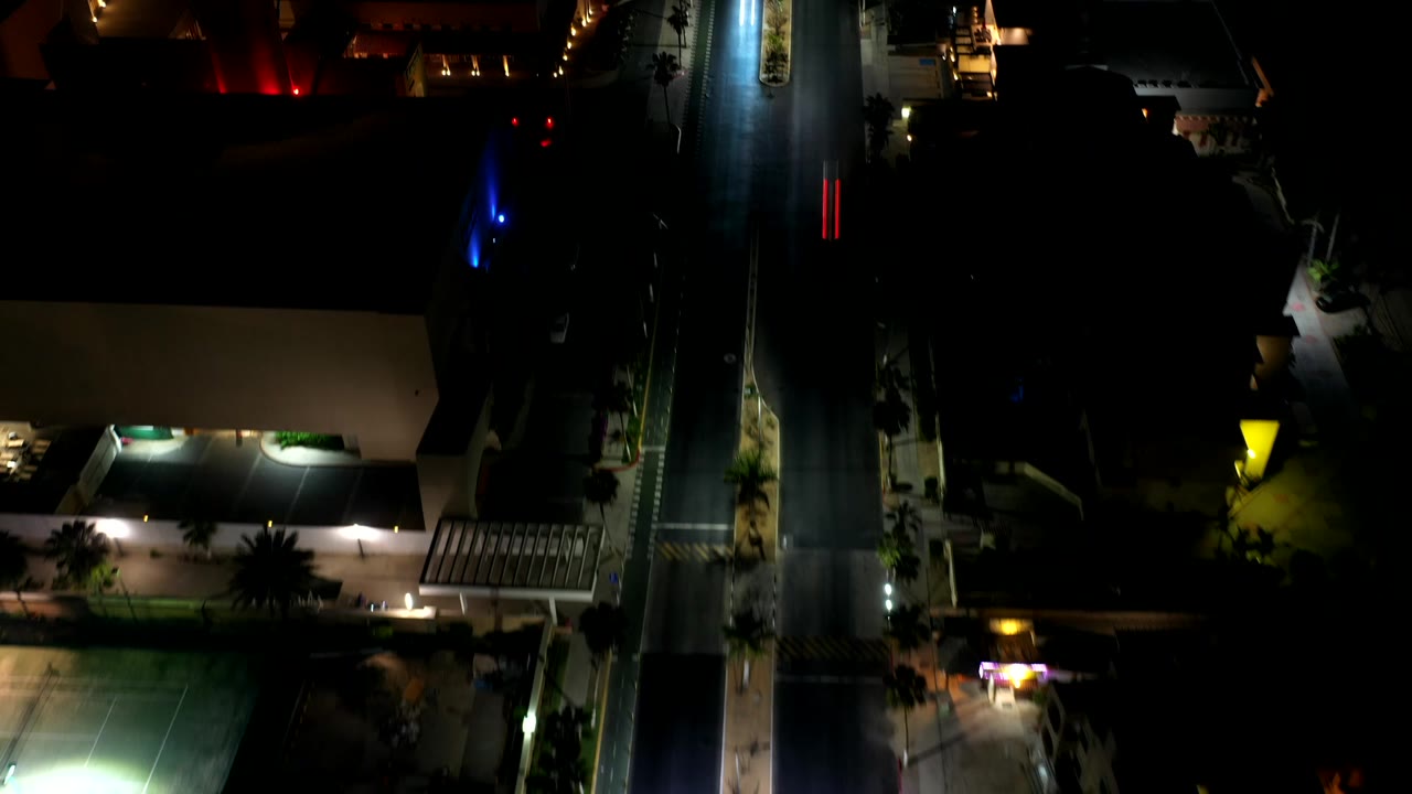 Opening overhead frame in Los Cabos shows a vertical road between dark buildings, warm roof lights, small colored light streaks, and no visible people.