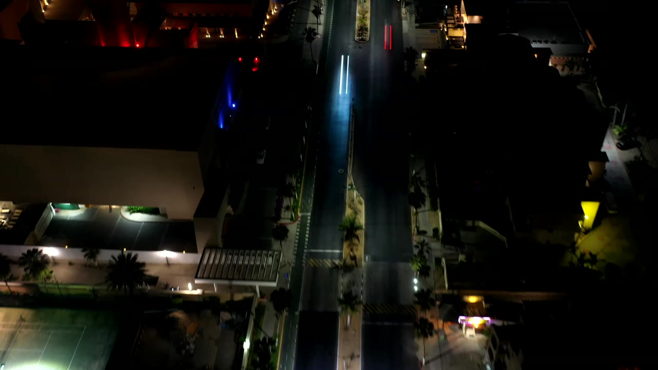 Los Cabos night aerial frame shows the road centered vertically, a lit building edge at lower left, dark roof areas, and no visible people.