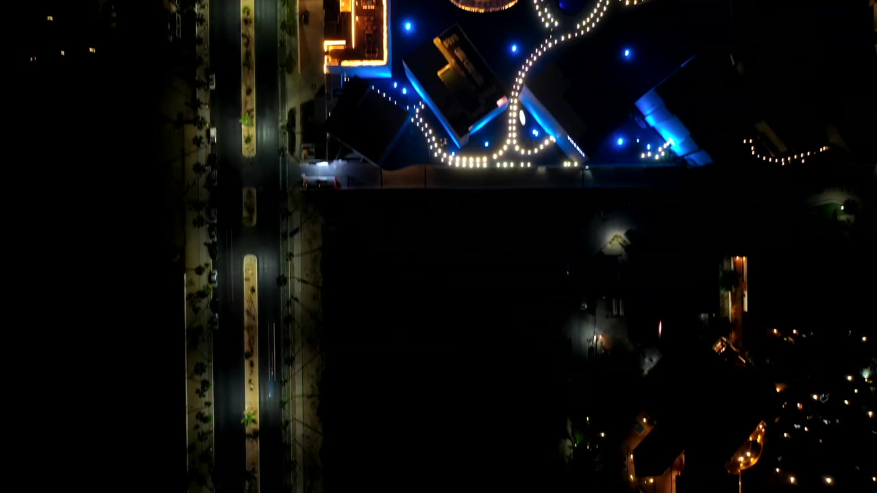Los Cabos night aerial frame with a vertical road along the left side, bright building edges near the top, curved white lights, and no visible people.