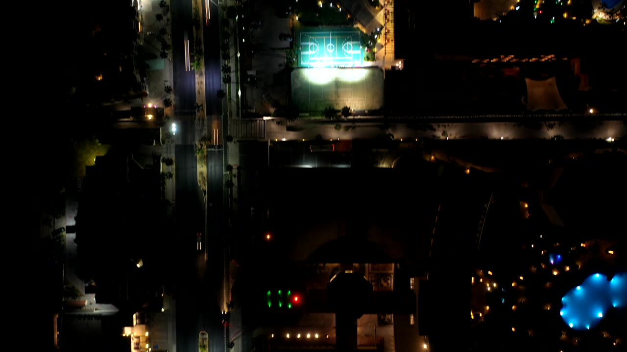 Overhead night frame in Los Cabos, Mexico showing a bright turquoise sports court near roads, dark rooftops, scattered streetlights, and no visible people.