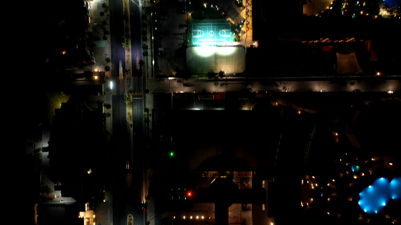 Overhead Los Cabos night view with the turquoise court near the top, warm road lights, a blue pool-like illuminated area, dark roofs, and no visible people.