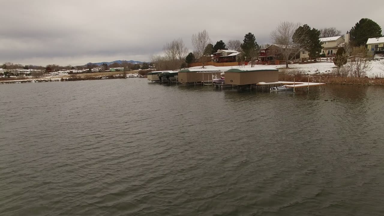 Snowy lakeshore homes and docks across rippled water under gray clouds.
