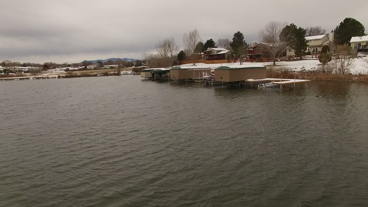 Second frame of the Jefferson County, Colorado lakefront, showing choppy water in front of snow-covered docks, small shoreline structures, houses, trees, distant hills, and no visible people.