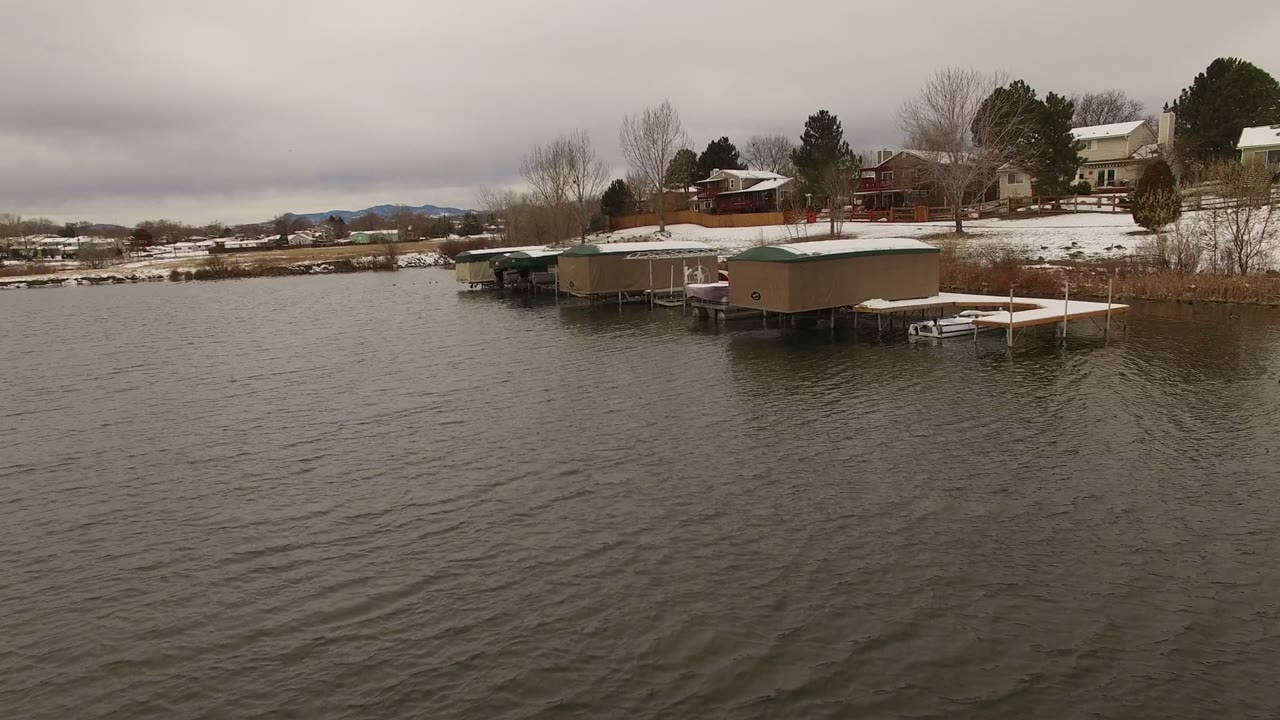 Snowy lakeshore with covered docks and houses beyond rippled water.
