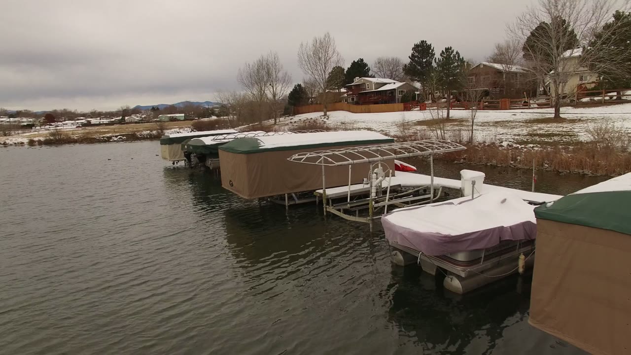 A low aerial view in Jefferson County, Colorado shows tan and green boat covers near metal docks, snow along the bank, winter trees, background houses, and no visible people.