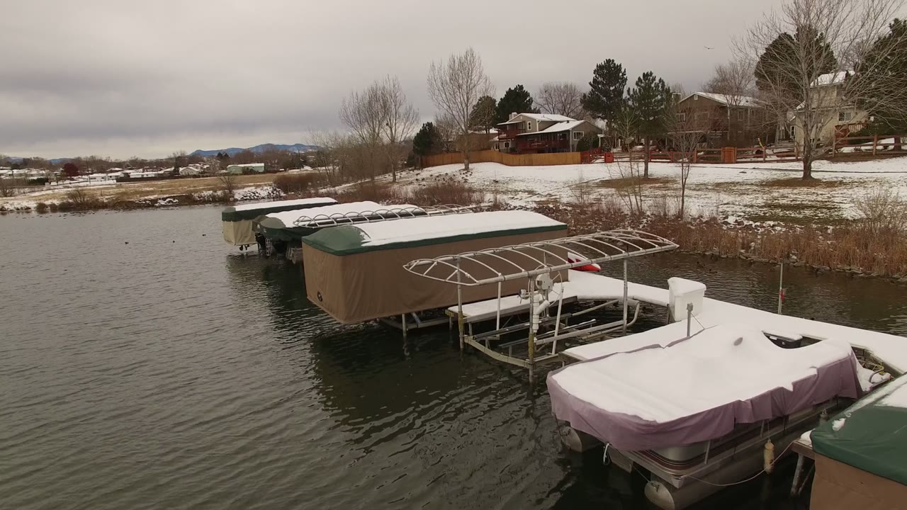 Aerial view in Jefferson County, Colorado of covered docks and boats edged by dark lake water, snowy yards, bare trees, distant hills, and no visible people.