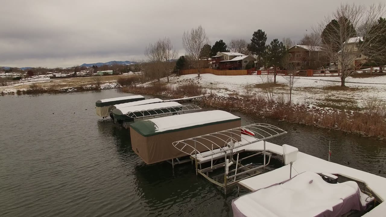Drone-height frame in Jefferson County, Colorado looking over multiple covered docks, rippled lake water, snow patches on yards, bare shoreline trees, and no visible people.