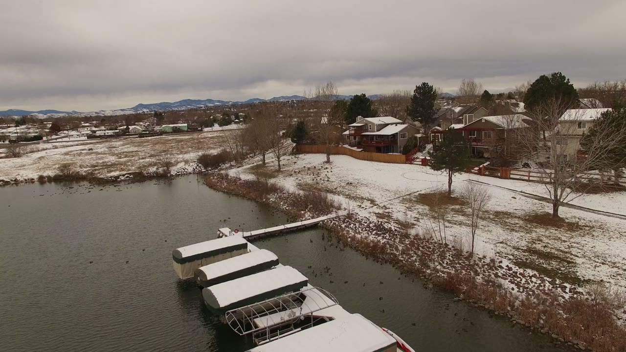 Snowy lakeside neighborhood with docks, houses, bare trees, and distant mountains.