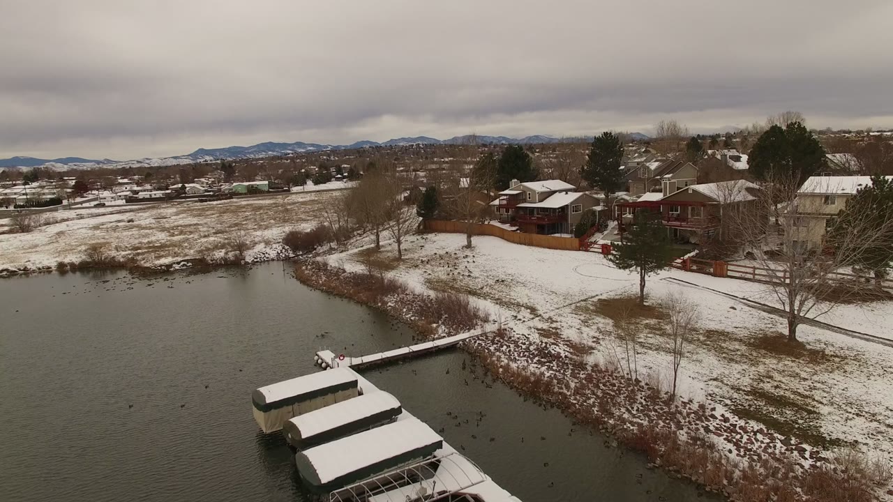 Aerial frame over a Jefferson County, Colorado lake edge, with white snow on dock roofs, brown winter brush along the shoreline, houses behind the water, distant mountains, and no visible people.