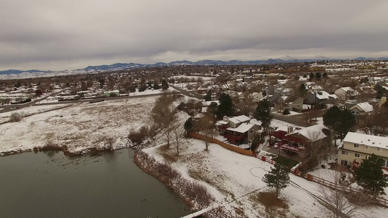 Elevated view of Jefferson County, Colorado homes, snowy roofs, bare trees, a lake edge in the foreground, and distant mountains under low gray clouds, with no clearly visible people.