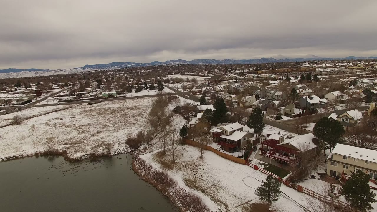 Jefferson County, Colorado winter aerial with a lake shoreline at lower left, fenced backyards, snow-dusted houses, leafless trees, and mountains across the horizon, with no clearly visible people.