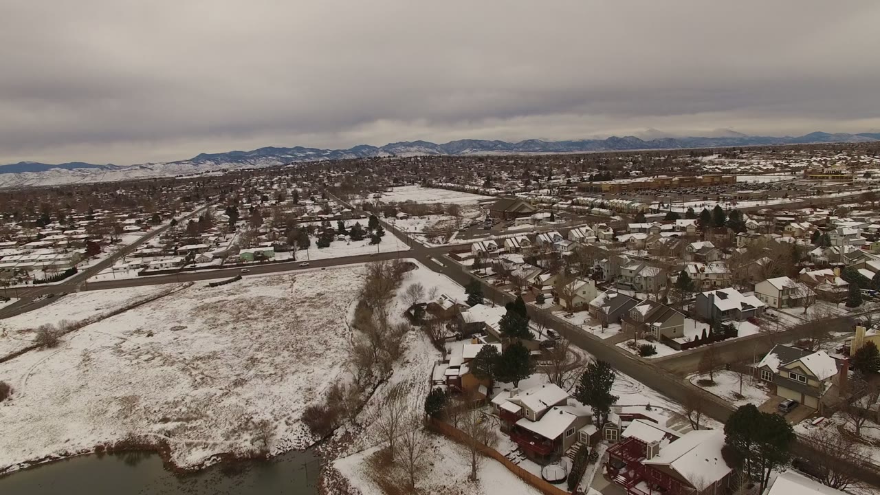 Aerial view of snow-covered suburban houses, roads, open field, and distant mountains under gray clouds.