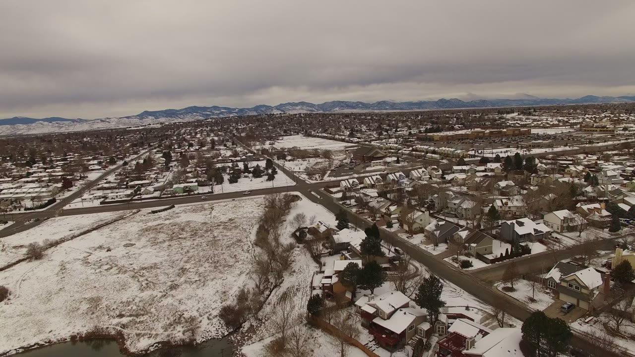 One second in, the Jefferson County, Colorado neighborhood remains spread below with more rooftops near the lower frame, a snowy field to the side, distant mountains, and no visible people.