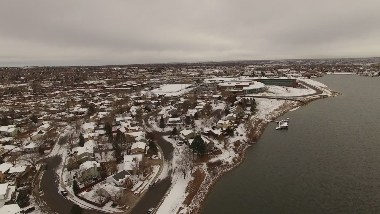 Aerial view of snow-covered lakeside homes beside dark water under gray clouds.