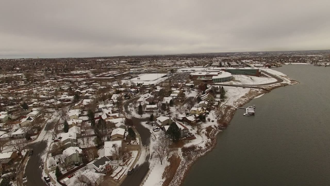 Aerial view in Jefferson, Colorado, with the lake occupying the right side, snow-dusted homes and streets on the left, small docks near shore, and no visible people.