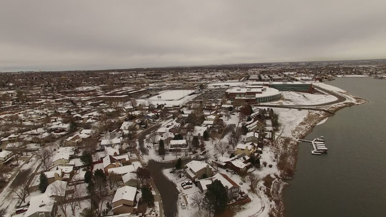 Wide aerial view in Jefferson County, Colorado showing snow-covered homes along a lakeshore, docks on the right edge, distant buildings, and no visible people.