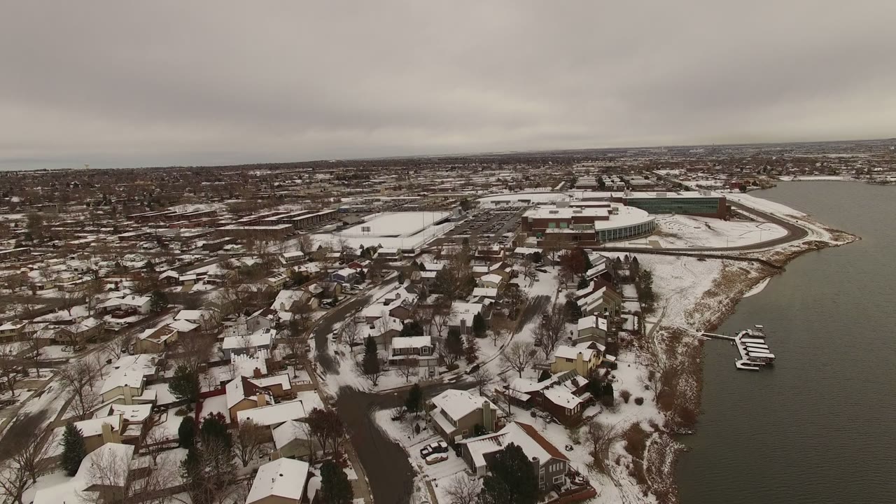 Jefferson County, Colorado aerial frame with clustered snowy roofs in the foreground, a curved lakeshore, small docks on the right, larger buildings beyond, and no visible people.