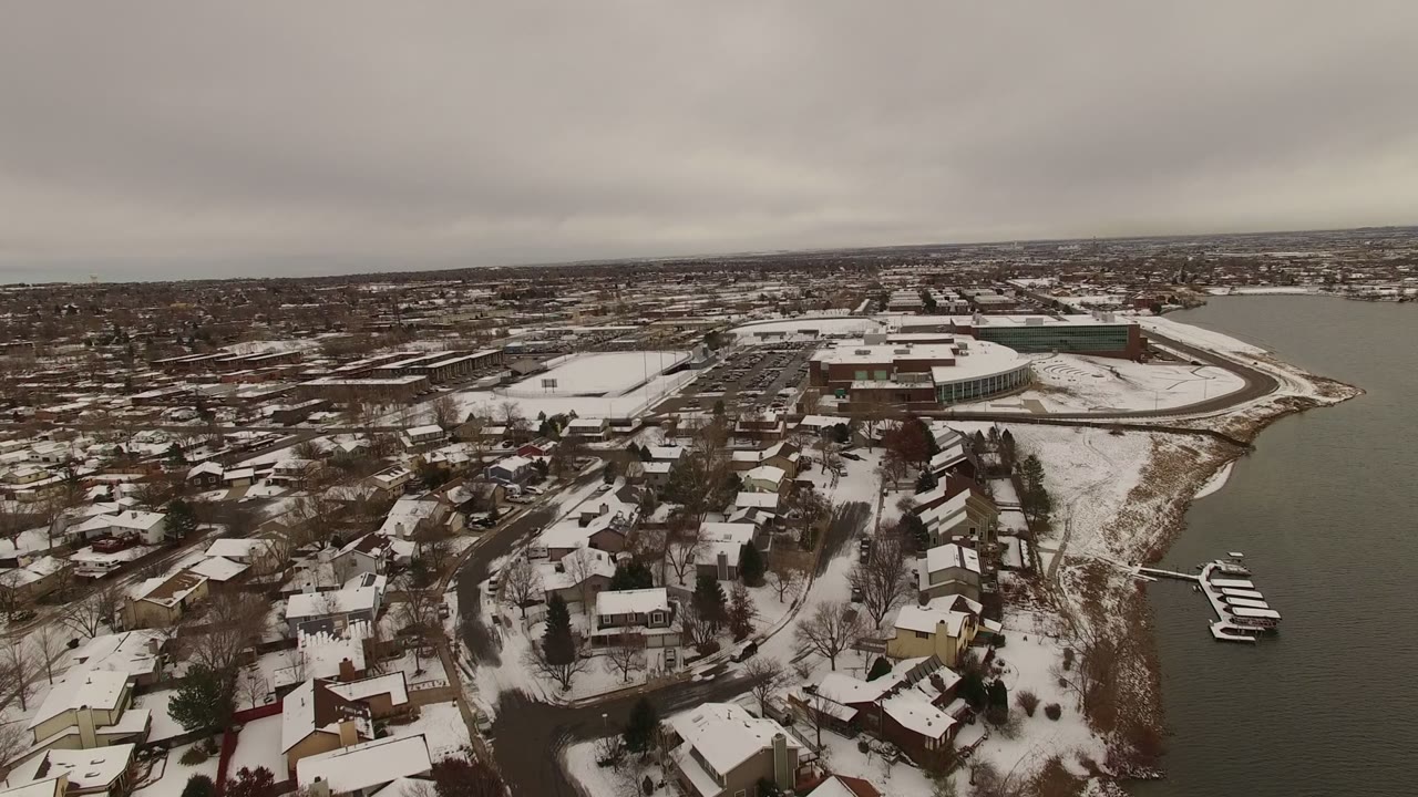 High aerial frame of Jefferson County, Colorado with clustered snowy rooftops in the foreground, docks and small boats near the shoreline, and no visible people.