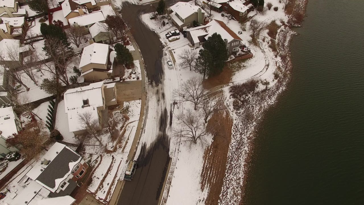 Snowy lakeside road with homes and dark water from above.