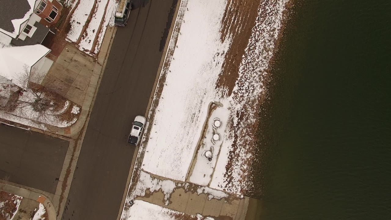 Overhead view of snowy shoreline beside a road and dark water.