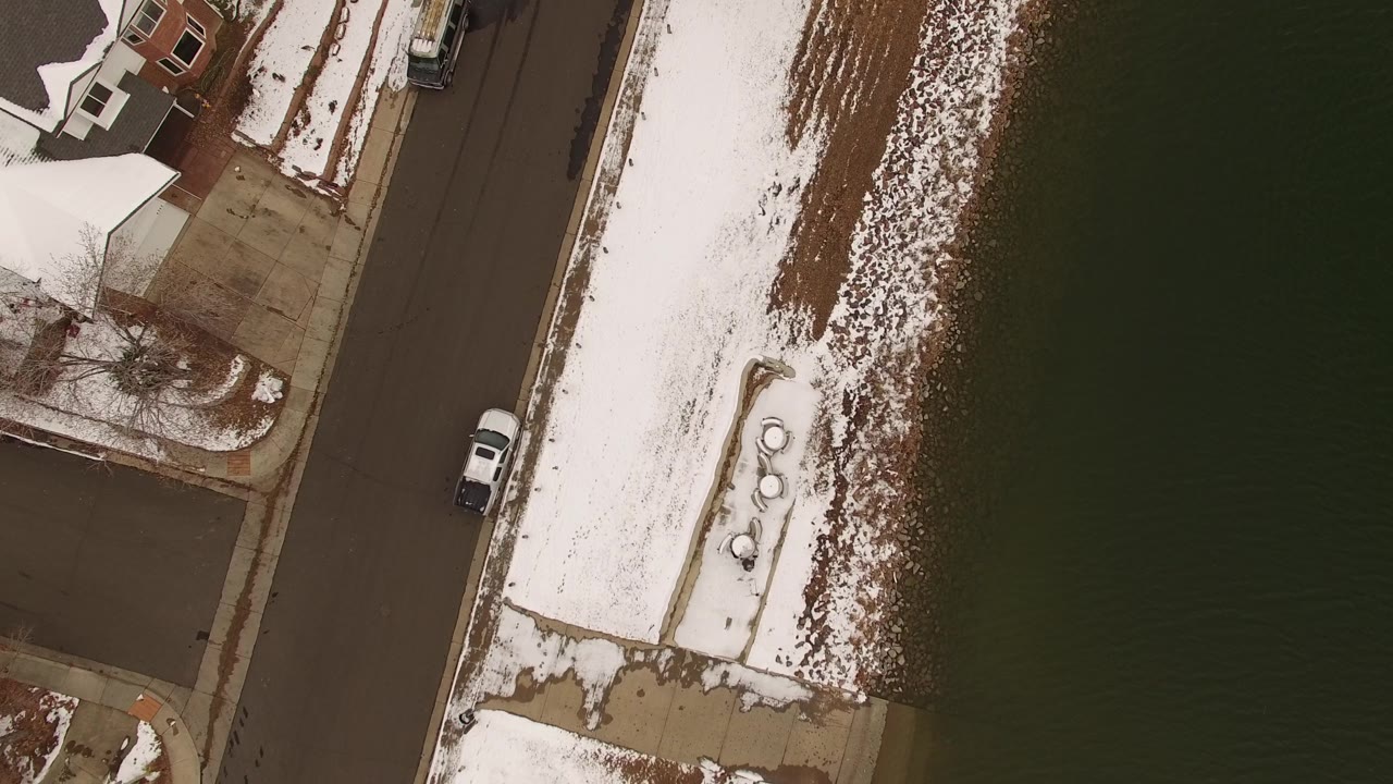 Second overhead frame in Jefferson, Colorado with the white vehicle lower on the road and the snow-covered shoreline centered beside dark water, with no visible people.