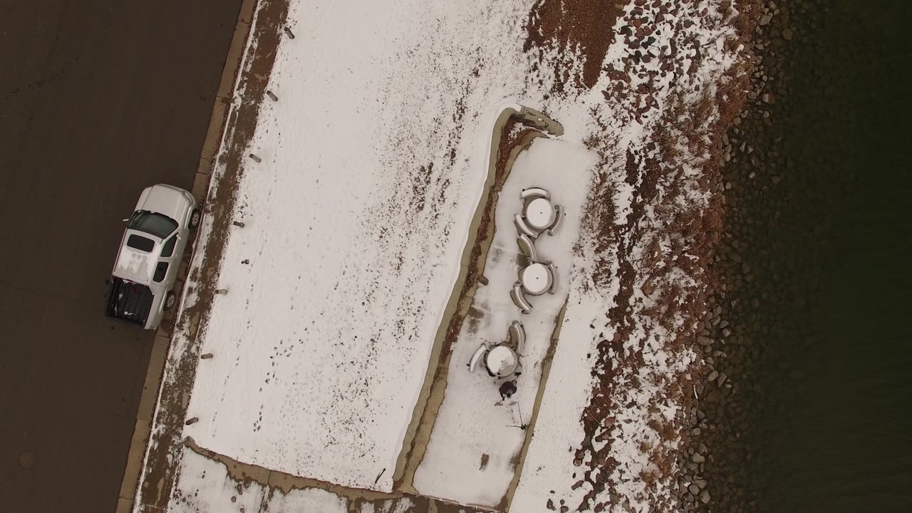 Top-down aerial frame in Jefferson County, Colorado, showing a white pickup along the left roadway edge, snow-covered grass in the center, and no visible people.