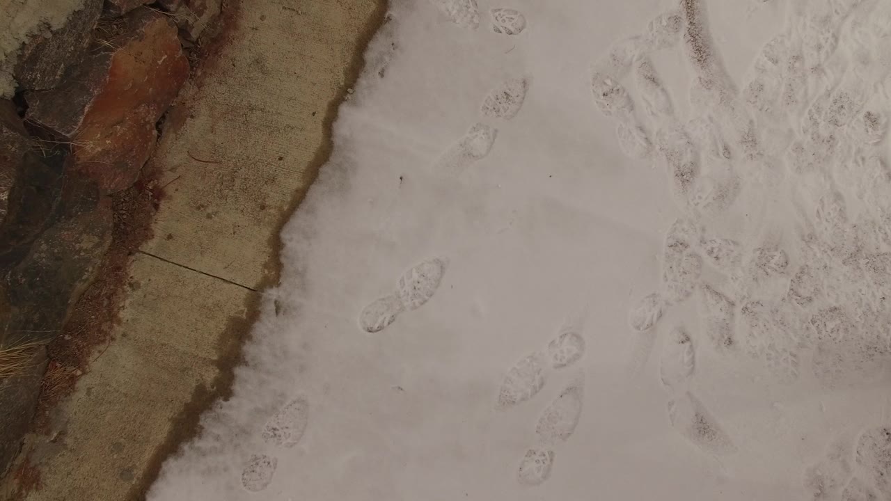 Overhead view of snowy ground in Jefferson, Colorado, with clustered footprints near a brown exposed ground edge and no visible people.