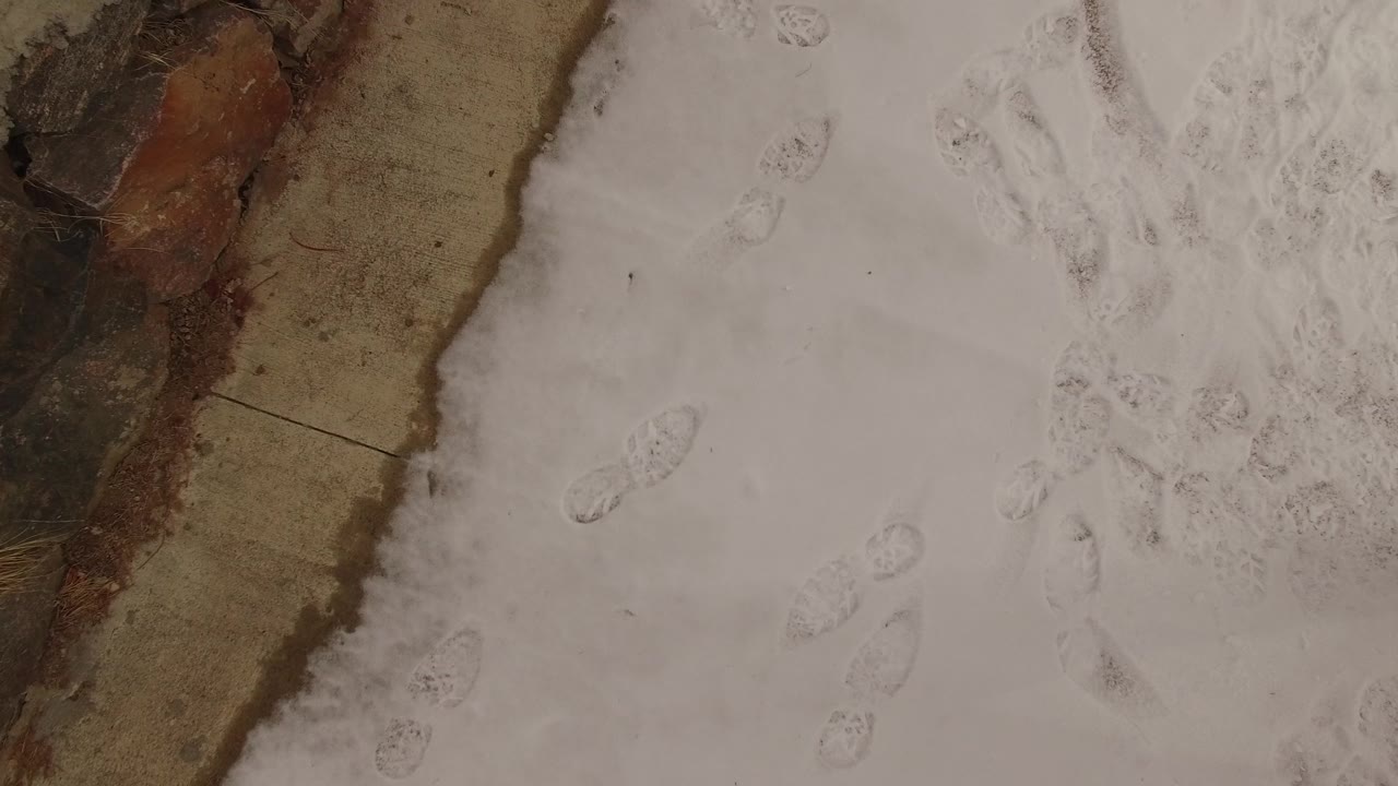 A closer overhead frame in Jefferson, Colorado shows pale snow marked by footprints beside a narrow tan and brown ground strip, with no visible people.