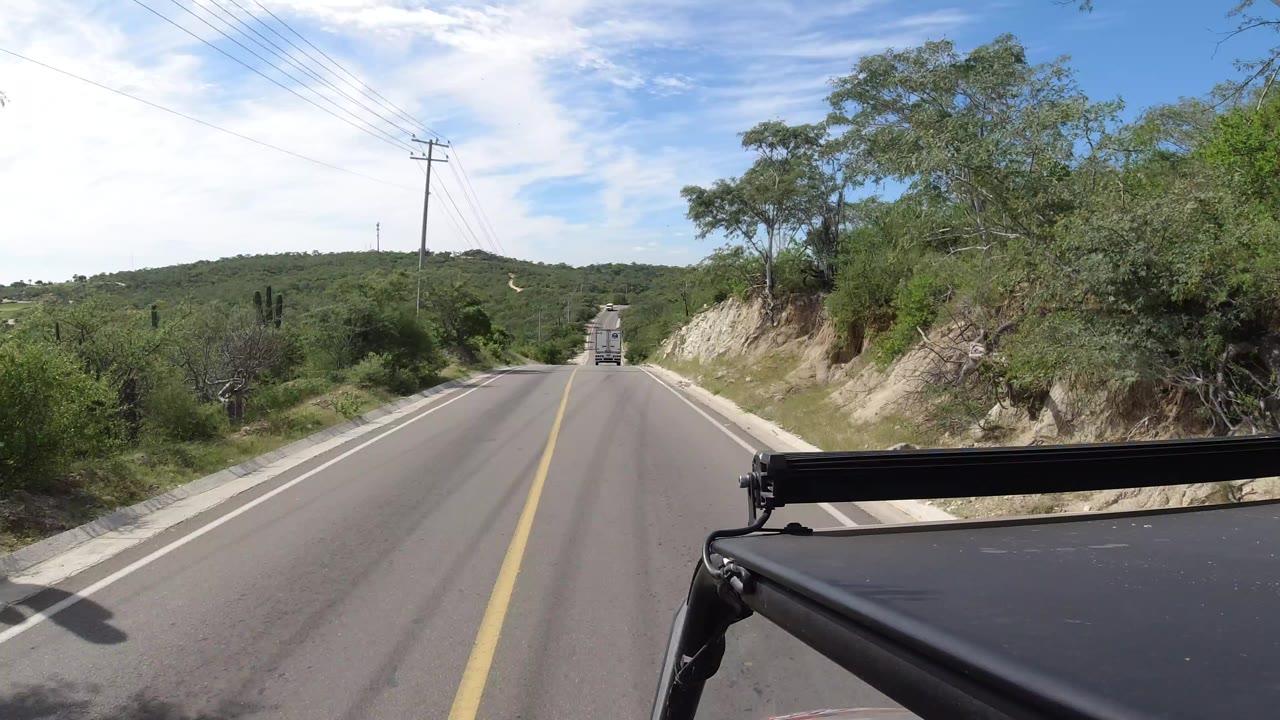 Car-top view in Los Cabos, Mexico looking down a two-lane road with utility lines, green hills, pale roadside rock, and no visible people.