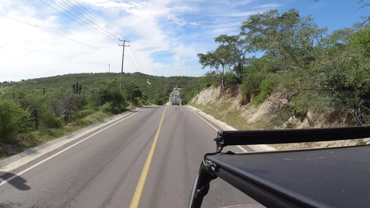 Forward-facing Los Cabos road view with the black car roof at lower right, a distant vehicle ahead, rocky shoulder, utility lines, and no visible people.