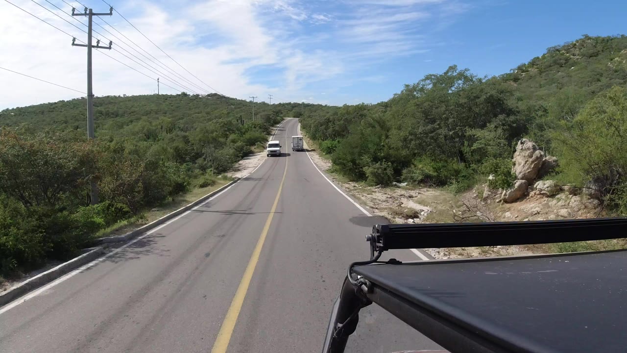Two-lane road through green brush with a white vehicle ahead