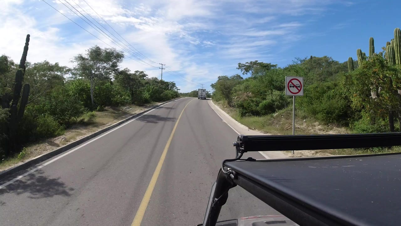 Start of a Los Cabos driving POV on a paved road bordered by cactus, scrub trees, sandy shoulder, overhead wires, and a no-passing sign, with no visible people.