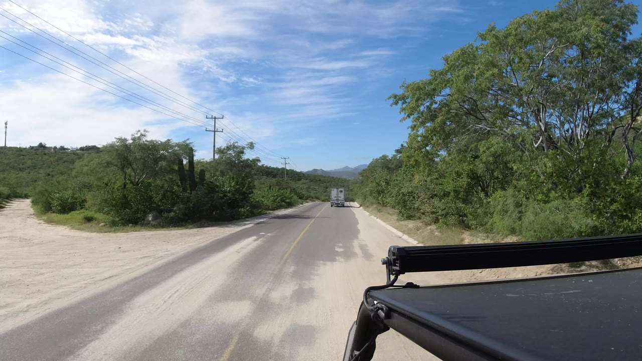 Forward road view in Los Cabos, Mexico, with a two-lane paved road, green trees on both sides, utility wires overhead, a vehicle ahead, and no visible people.