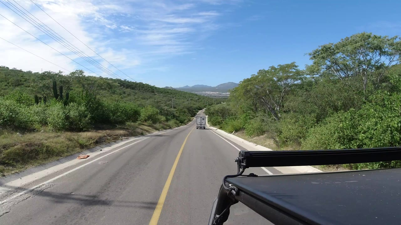 Opening frame of a Los Cabos road view with a white vehicle ahead, dense green brush on both sides, overhead power lines, distant hills, blue sky, and no visible people.