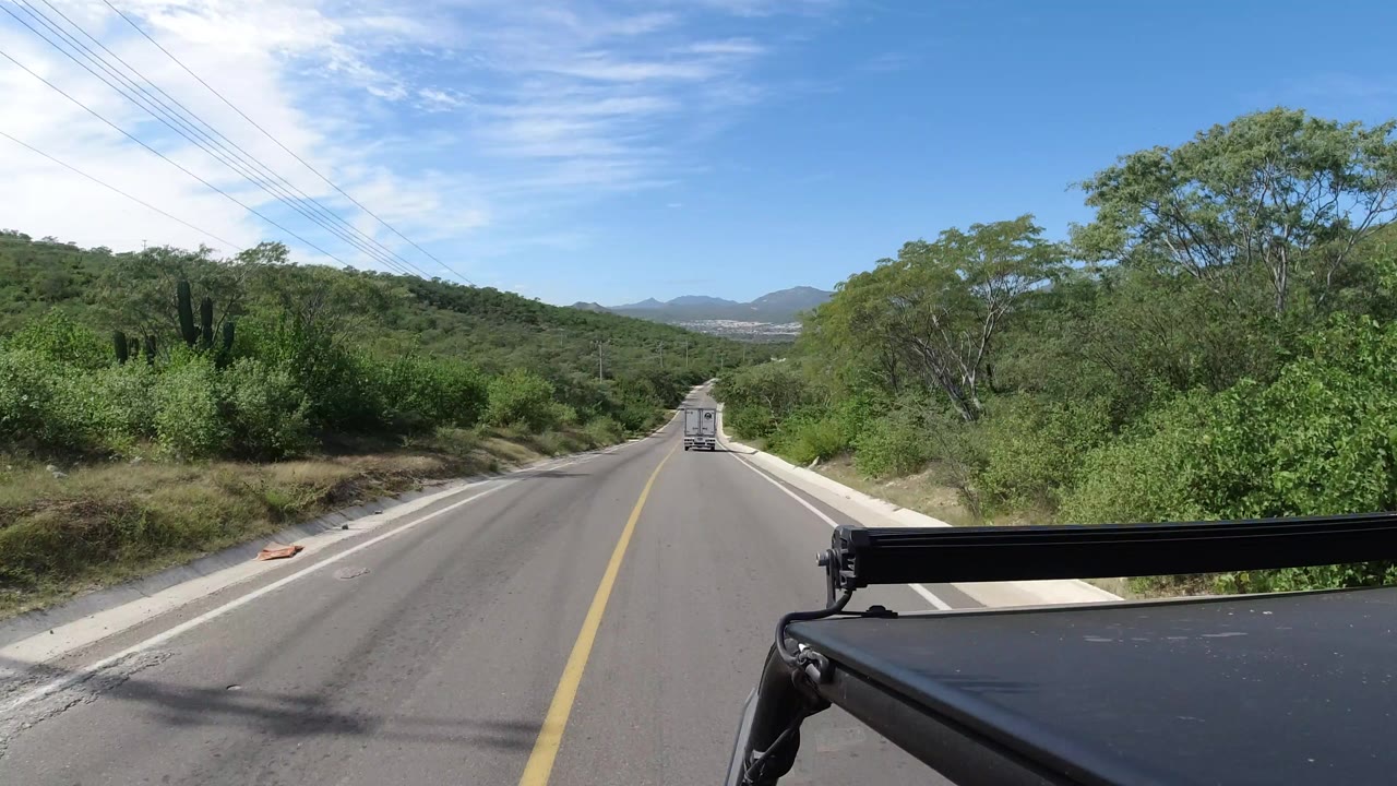 Los Cabos road scene one second in, with the centered yellow line leading toward a white vehicle, thick roadside vegetation, low hills, blue sky, and no visible people.