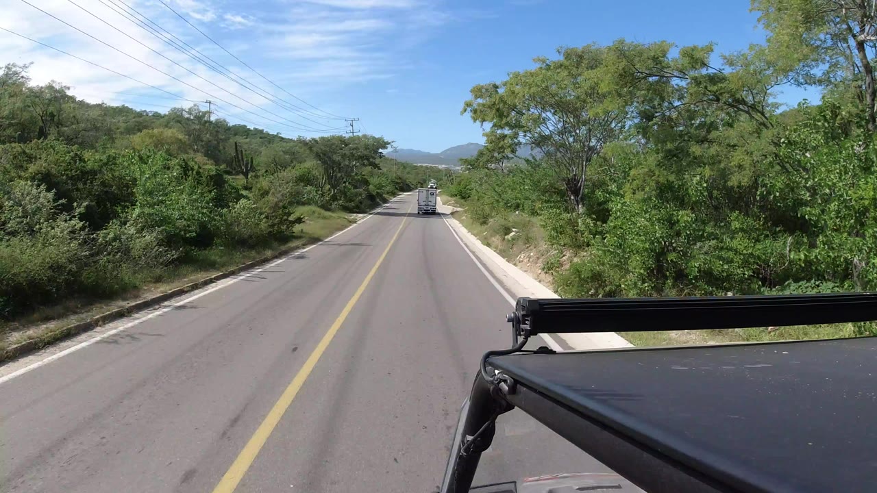 Forward road view in Los Cabos, Mexico, with green roadside vegetation, utility lines overhead, a distant vehicle ahead, part of the filming vehicle at lower right, and no visible people.