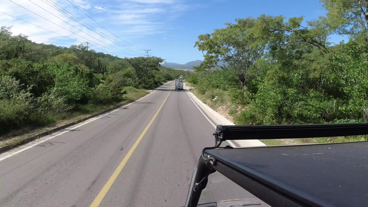 Los Cabos road view with a straight lane, dense green brush on both sides, distant hills on the horizon, vehicle hardware in the lower foreground, and no visible people.