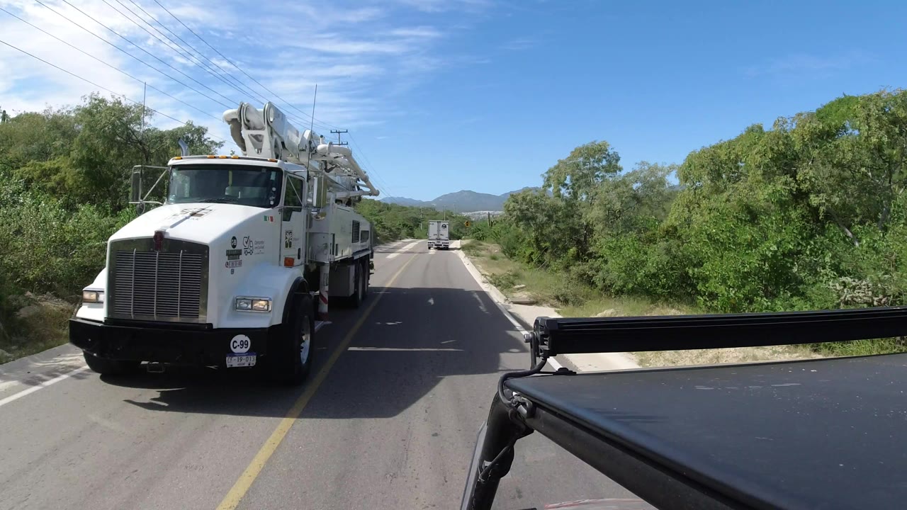 The white utility truck remains alongside the camera on the Los Cabos road, with a second vehicle farther ahead, roadside brush, overhead wires, and no visible people.