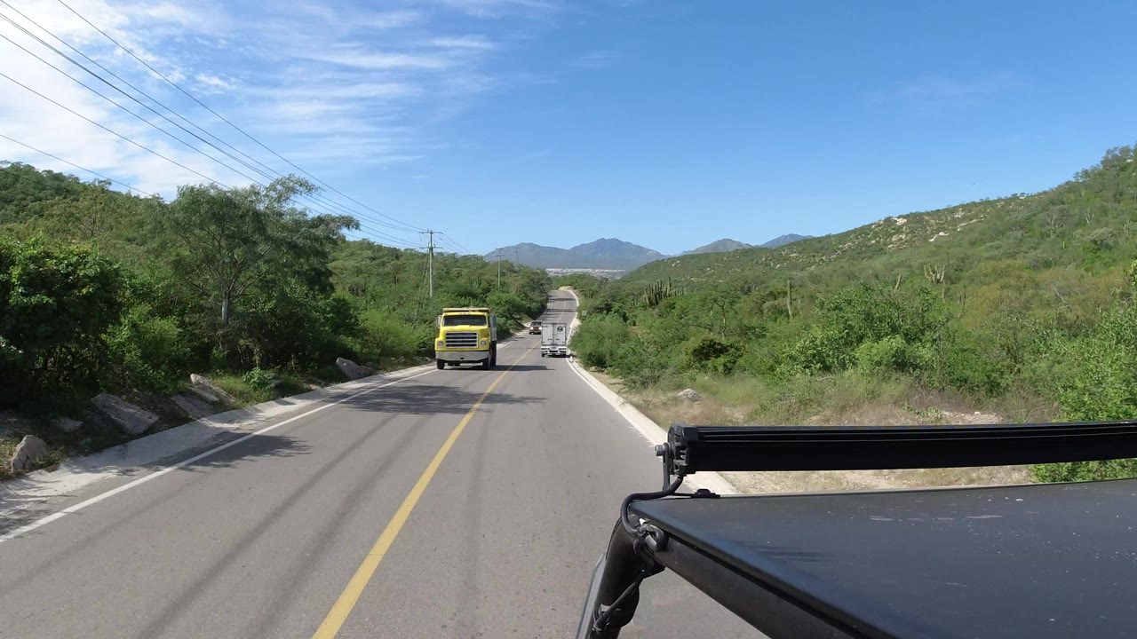 Los Cabos, Mexico road frame with the yellow truck farther down the lane, a white vehicle beyond it, low green hills, overhead utility lines, and no visible people.