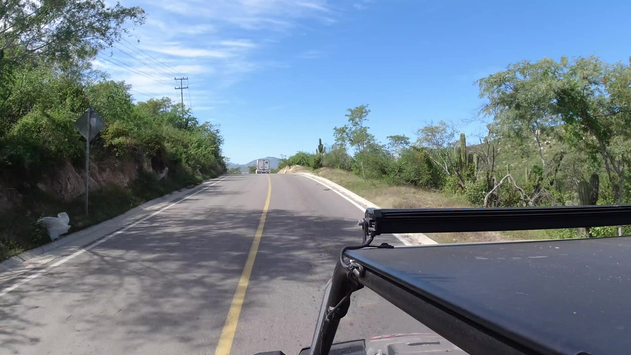 Opening frame of a Los Cabos, Mexico road seen from a vehicle, with a yellow centerline, green roadside brush, distant hills, utility poles, vehicle hardware in the foreground, and no visible people.
