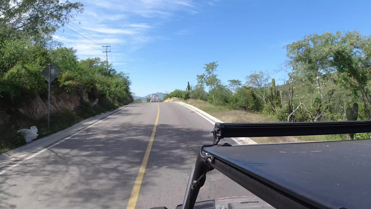 A later Los Cabos, Mexico road frame shows the vehicle continuing along the two-lane pavement, with dense green bushes on both shoulders, a utility pole ahead, distant hills, and no visible people.
