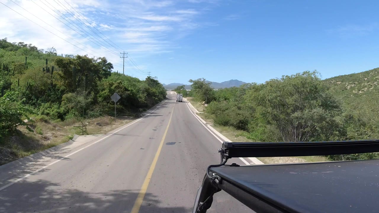 Forward vehicle view on a two-lane road in Los Cabos, Mexico, with green roadside vegetation, power lines at left, distant hills ahead, vehicle hardware at lower right, and no visible people.